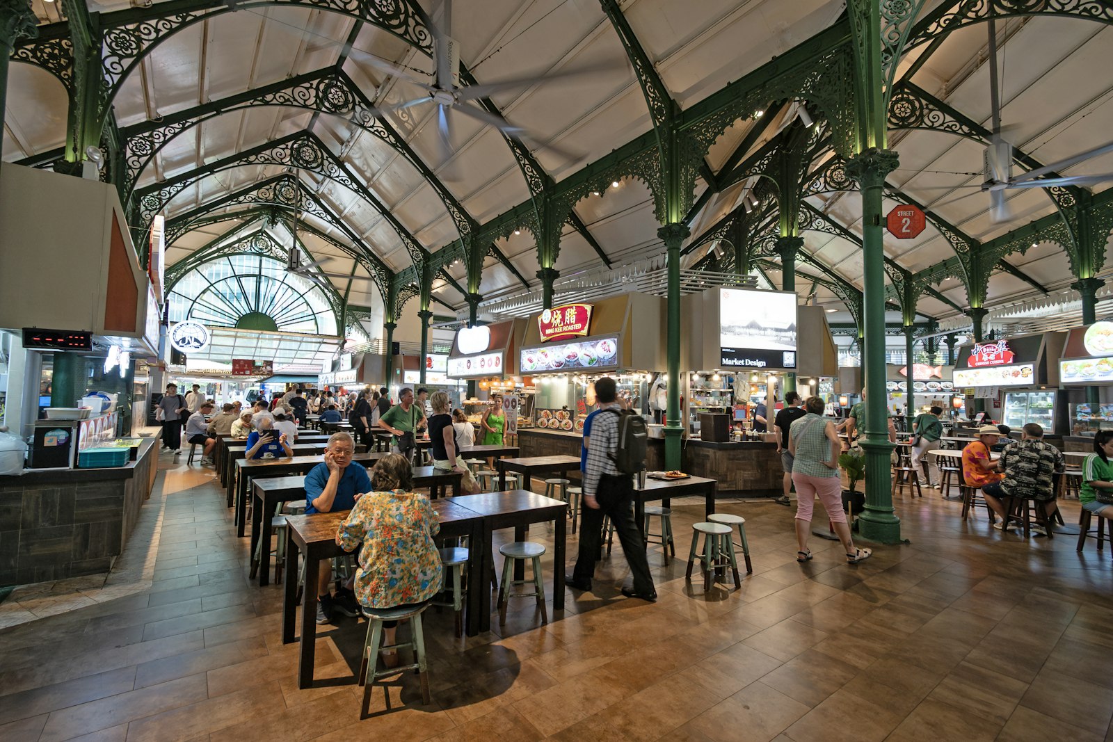 A group of people seated at communal tables inside Lau Pa Sat, a real Singapore hawker centre, Victorian ironwork and clock tower visible.