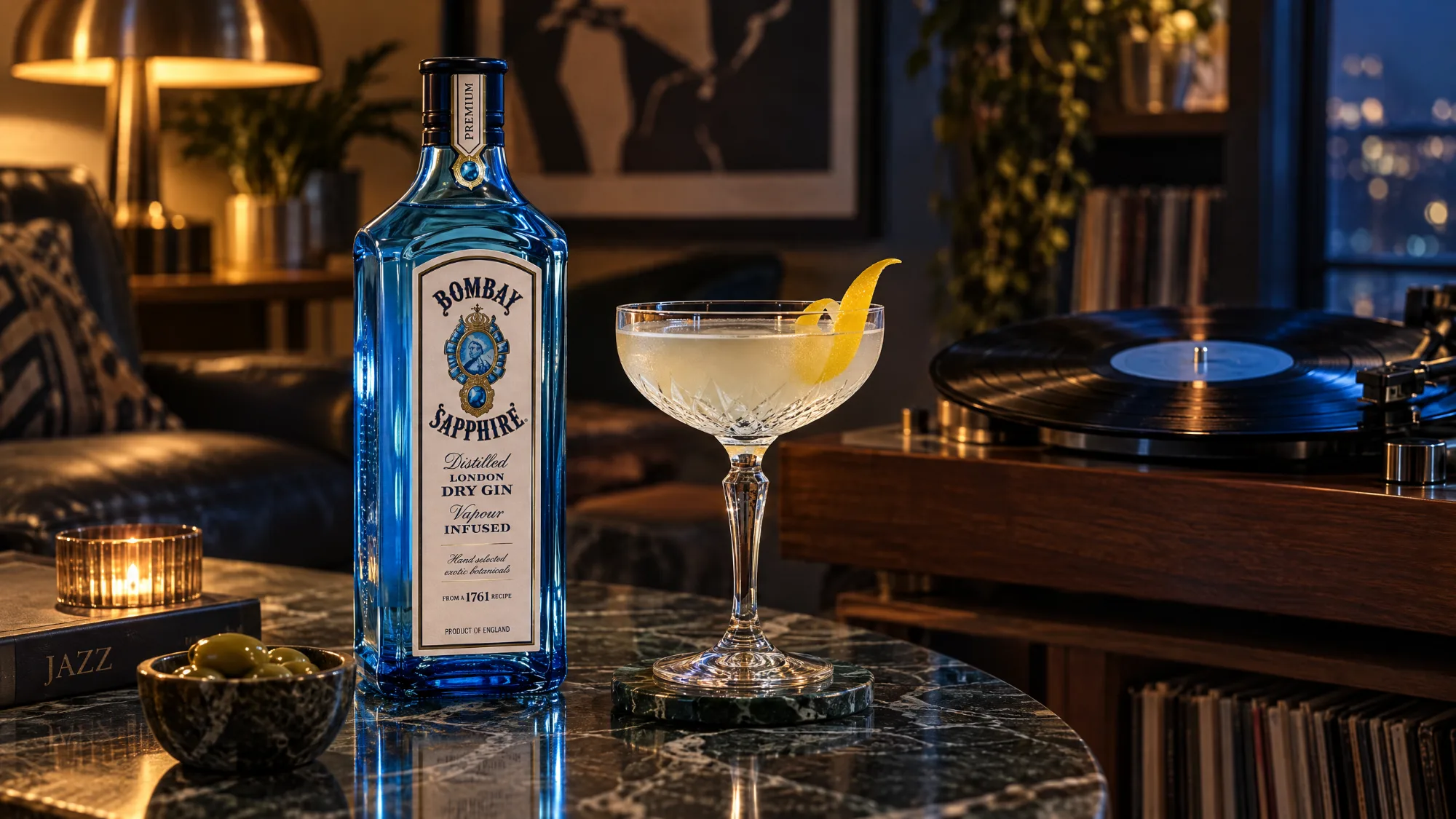 A blue bottle of Bombay Sapphire Gin beside a coupe glass with a lemon twist on a dark marble table, with a bowl of olives, a lit candle, a record player and shelves of records behind.